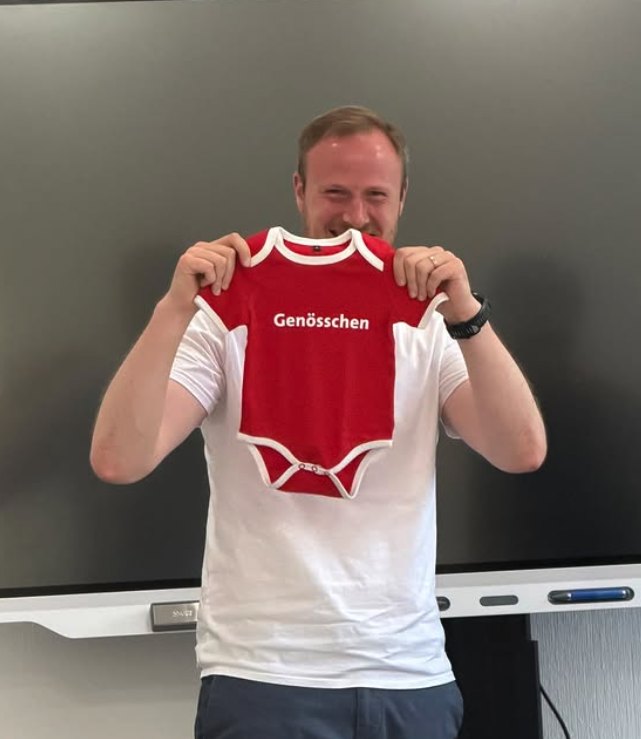 Philipp Wilp hält einen roten Babystrampler mit der Aufschrift „Genösschen“ hoch